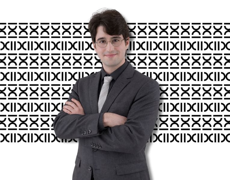 Advogado previdenciário especialista em planejamento previdenciário, trajando terno cinza escuro com gravata cinza claro, sobre o plano de fundo de padronagem com a logomarca do escritório.